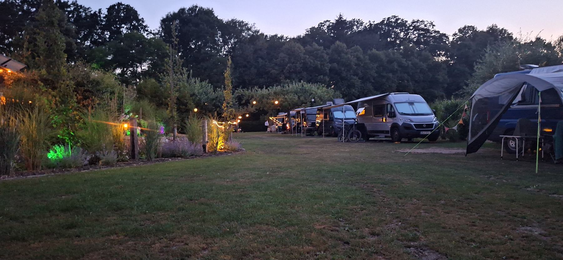 Der Campingplatz am Weberhof in der Abenddämmerung: Wohnmobile und Camper auf der Wiese, Lichterketten und beleuchtete Beete.